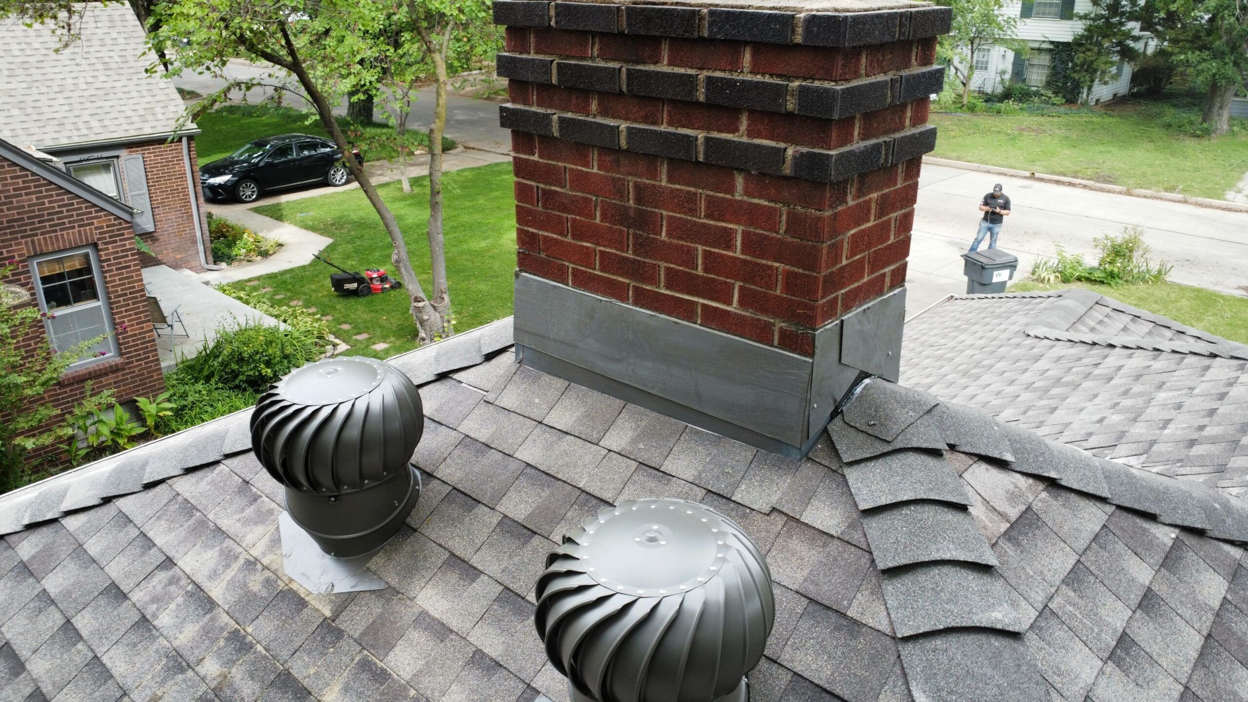 An asphalt shingle roof with a shallow pyramid hip roof and sufficient space for two wind turbines downslope from a brick chimney.