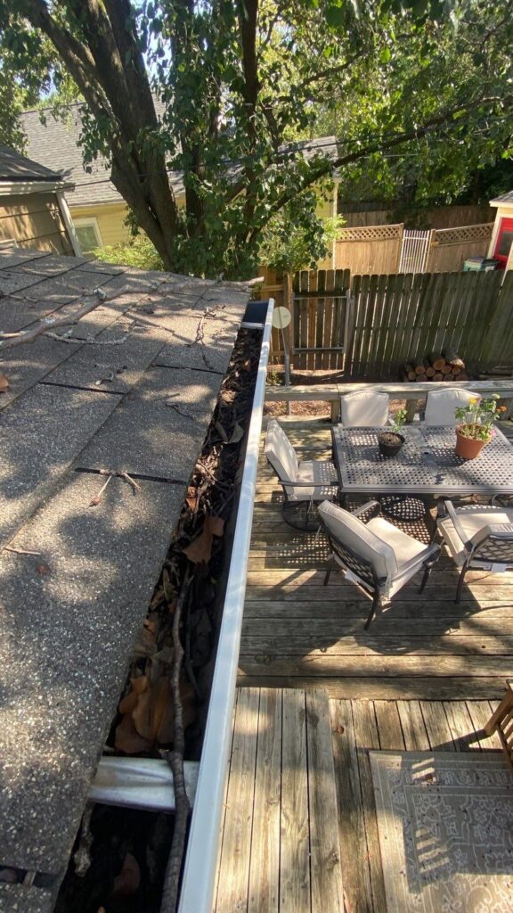Clogged gutters filled with leaves, twigs, and debris alongside a wooden deck with patio furniture.