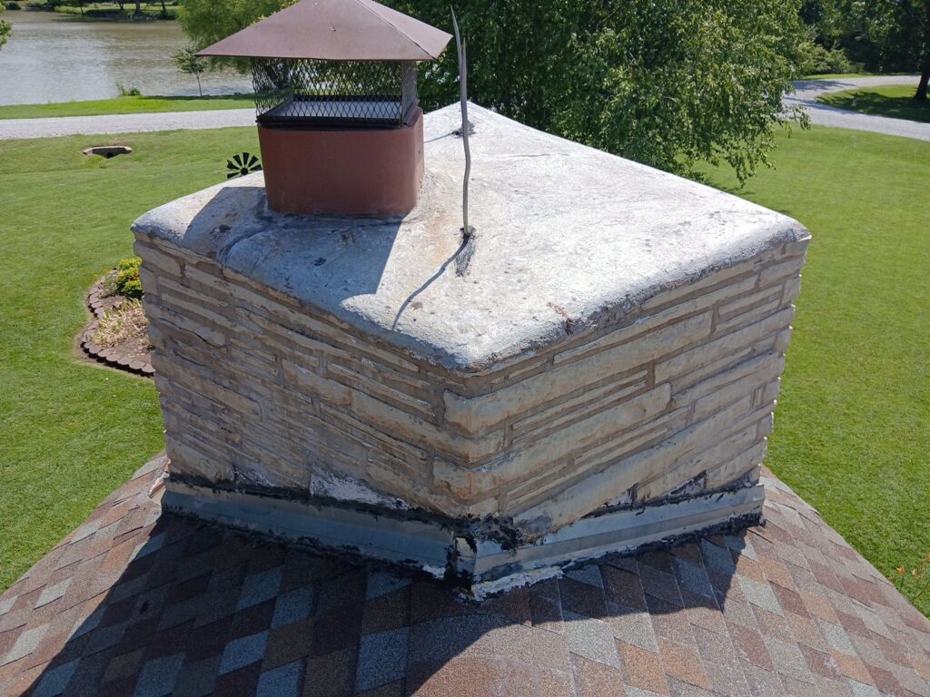 Surface-mounted counterflashing on a large stone chimney with previous repairs visible atop the counterflashing.