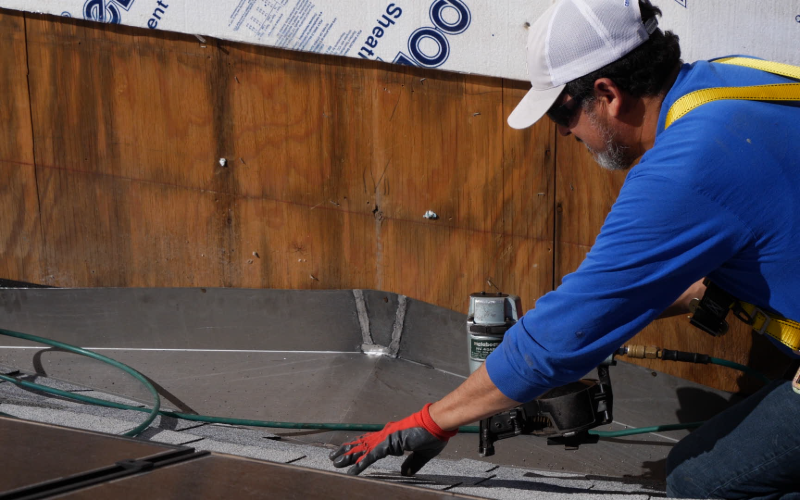 A worker wearing a safety harness and using a nail gun re-fastens chimneys abutting a metal chimney cricket on an asphalt shingle roof.