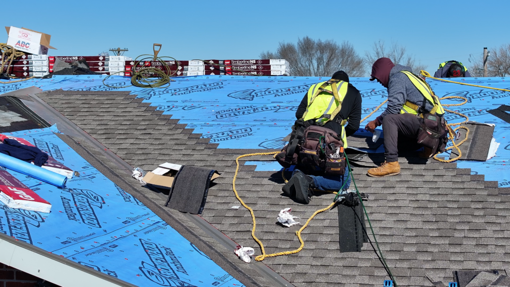 Two roofing crew members install shingles near a metal valley with underlayment below. 