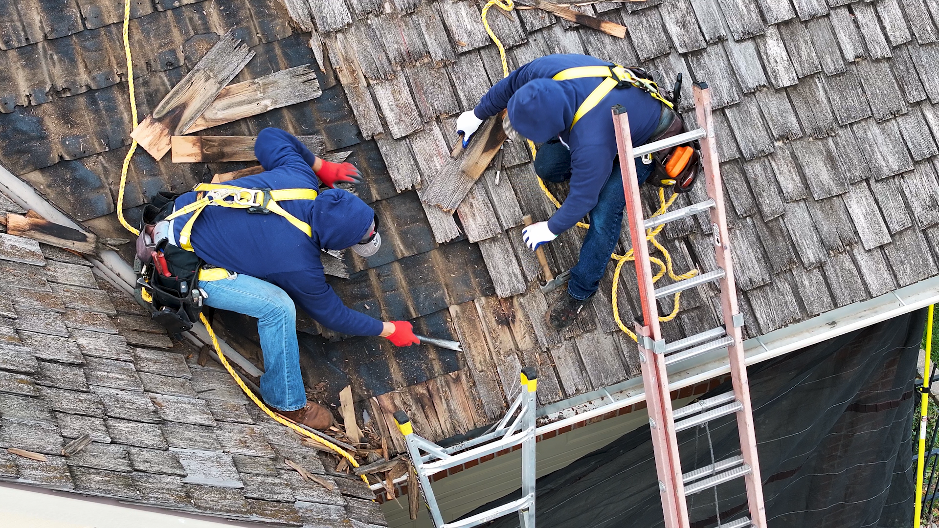 Two workers tearing off wood shake shingles with prybars during a residential roof replacement.