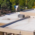 A low-slope commercial roof with parapet walls and coping caps on a TPO field with internal drains, lines on blocks, and an HVAC unit.