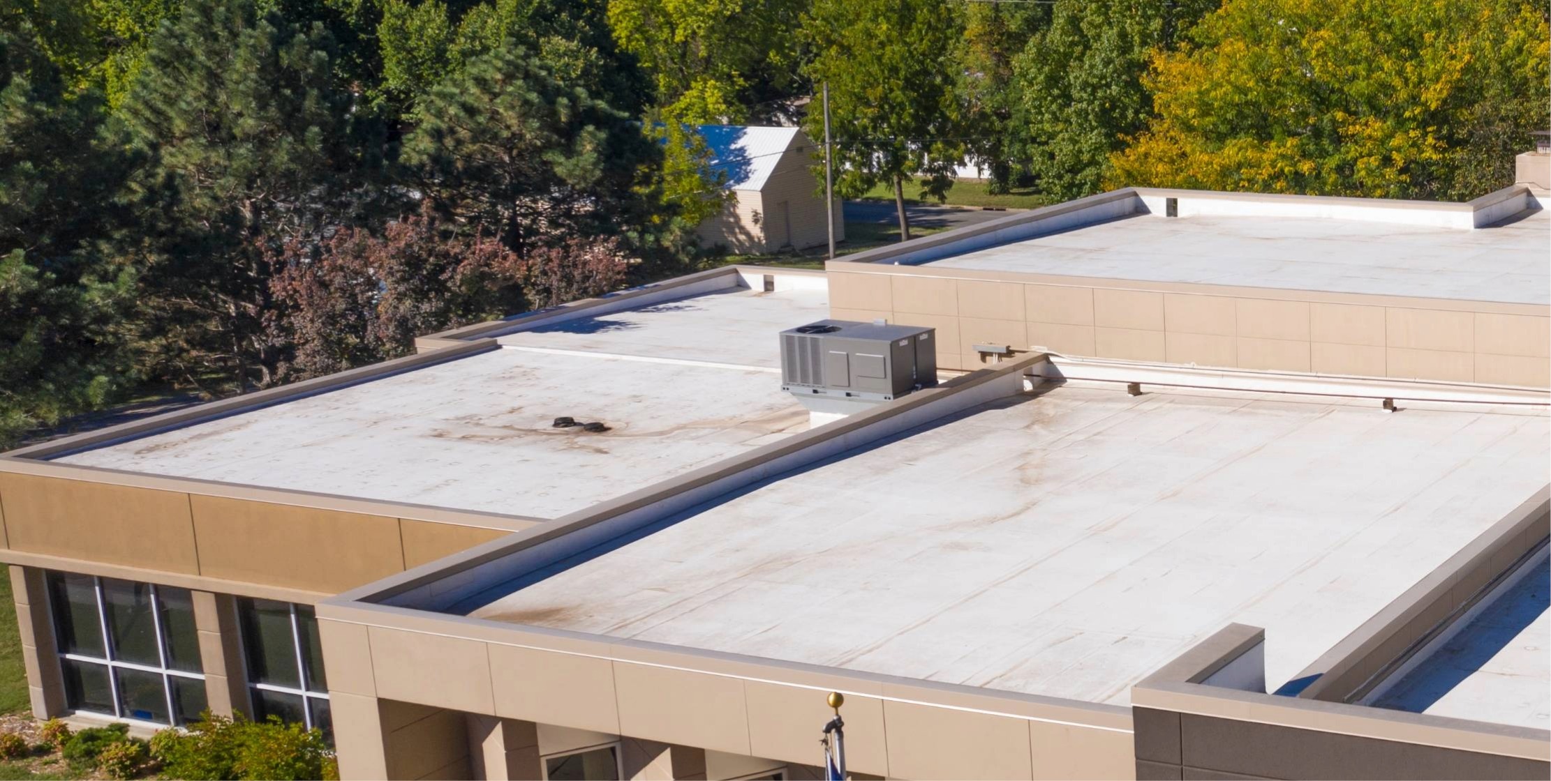 A low-slope commercial roof with parapet walls and coping caps on a TPO field with internal drains, lines on blocks, and an HVAC unit.