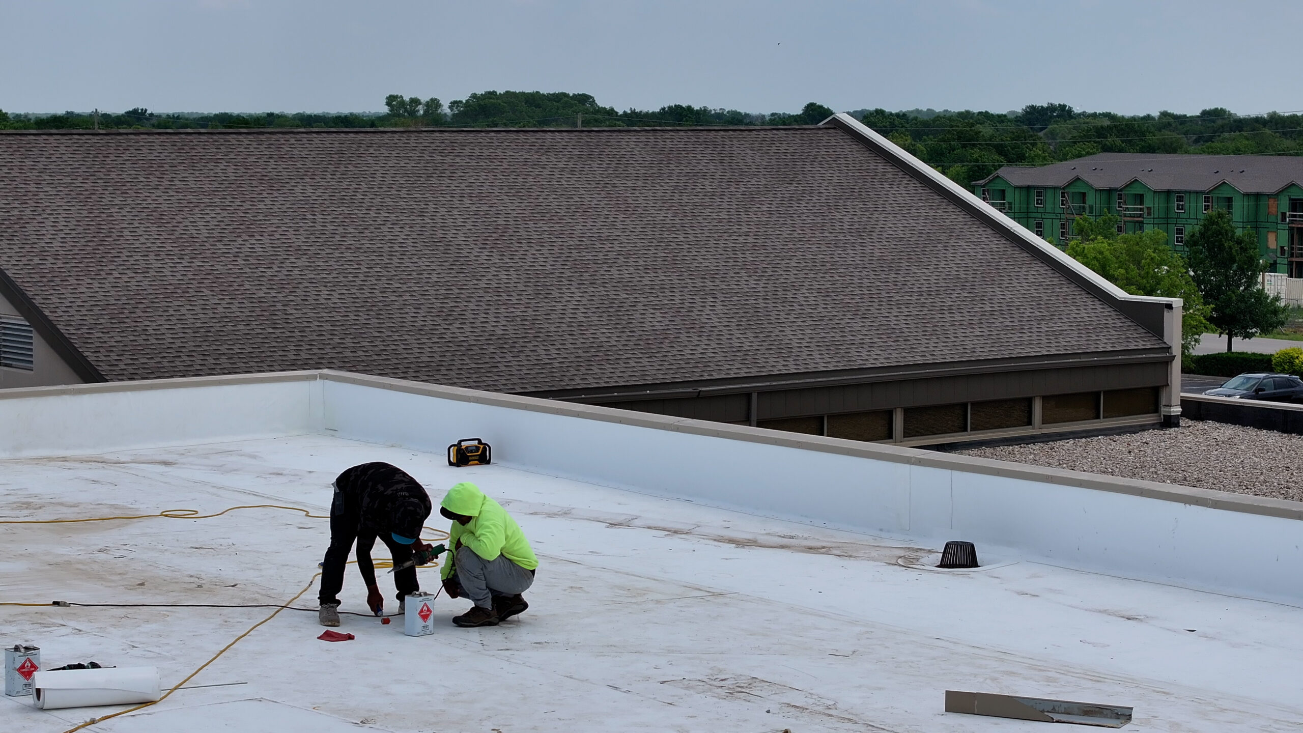 Two workers complete repairs on a new low-slope TPO roof system with a ballasted EPDM roof and an asphalt shingle roof system in the background.