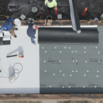 A single-ply membrane installation on a low-slope roof with cover board below and three roofers preparing material.