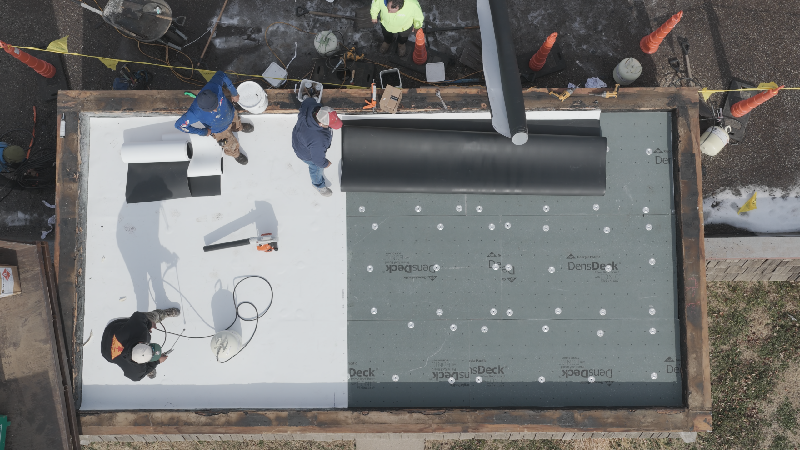 A single-ply membrane installation on a low-slope roof with cover board below and three roofers preparing material.