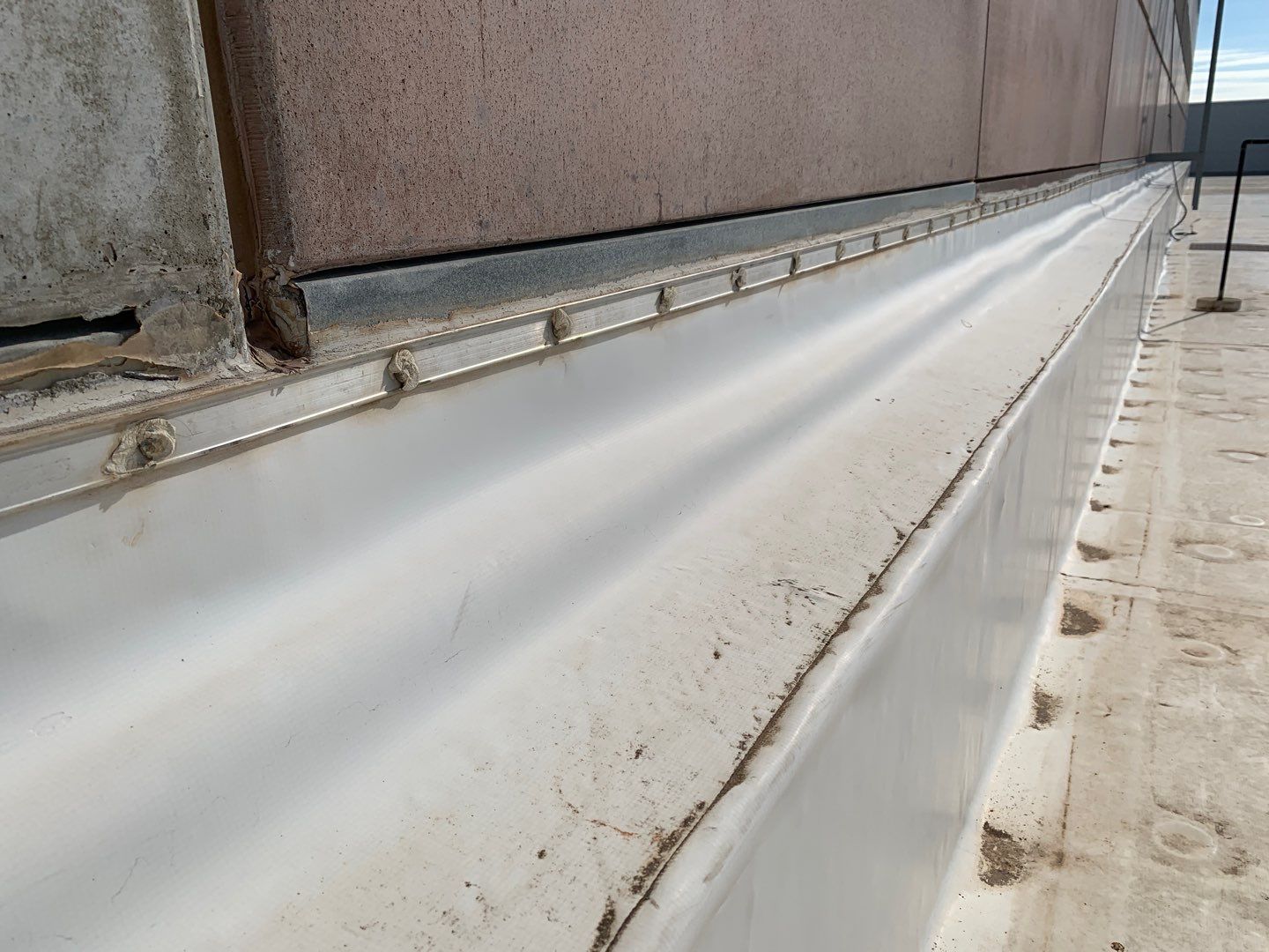 A termination bar on a white membrane roof sealing the cut membrane at a roof-to-wall transition.