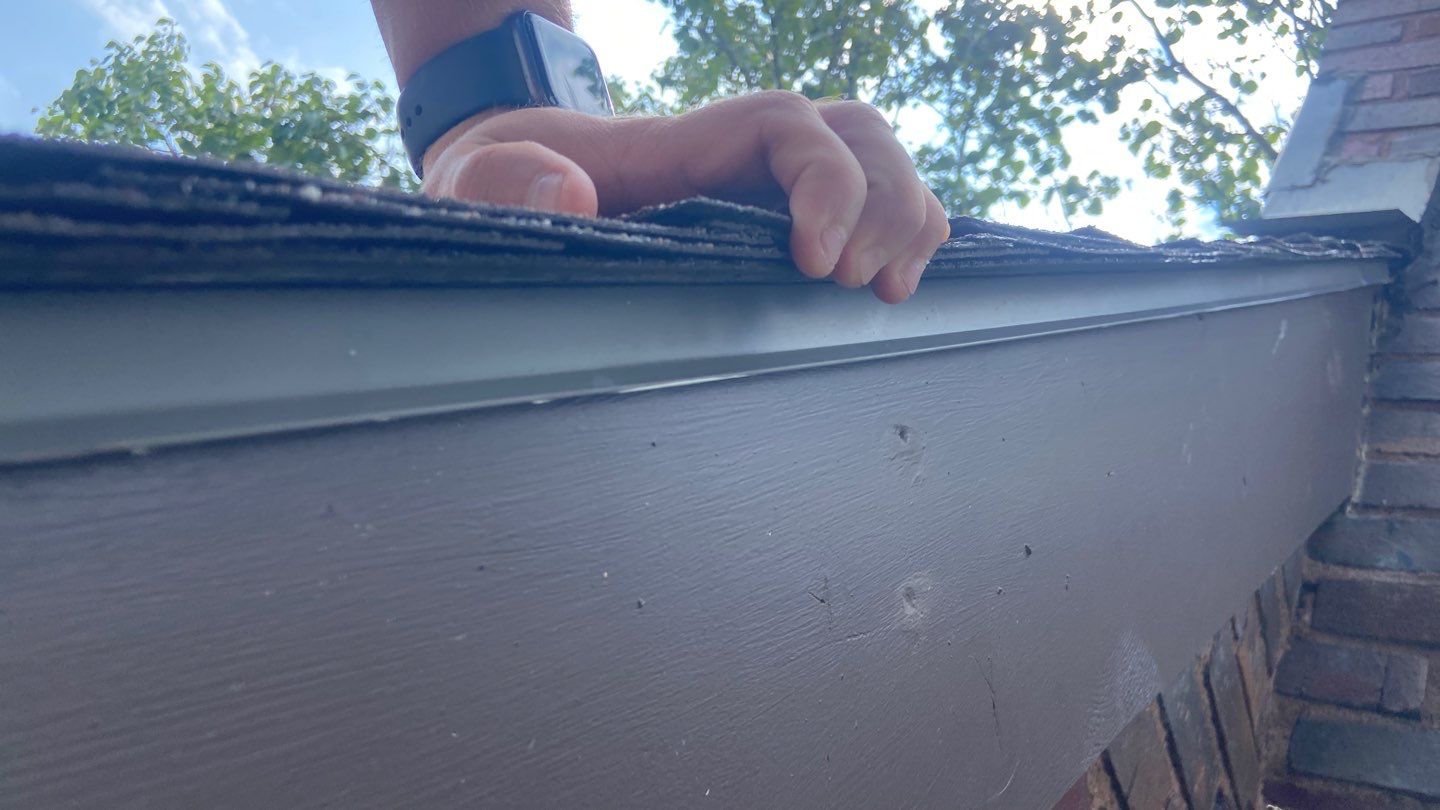 A human hand with a watch on an asphalt roof showing the drip edge at the rakes with a chimney and its flashing in the background.