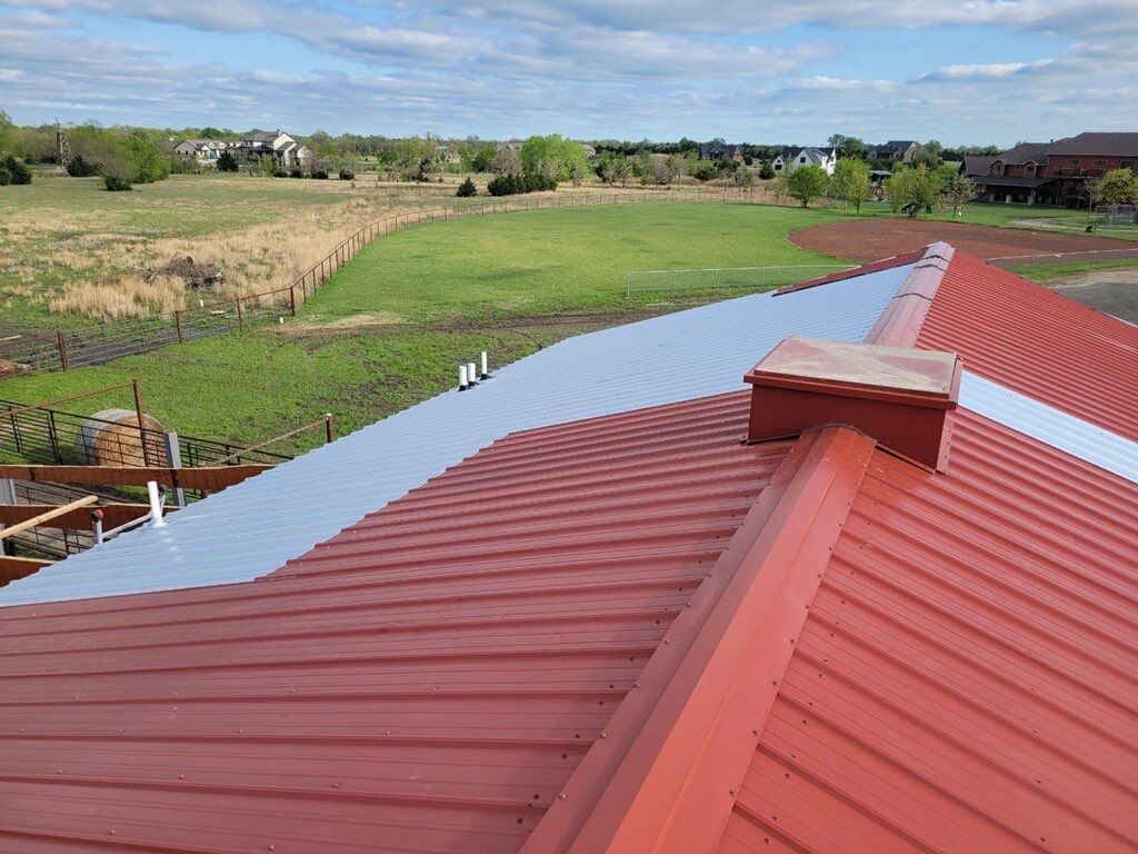 A red metal roof on a barn with unpainted metal repair areas and a baseball field in the background.